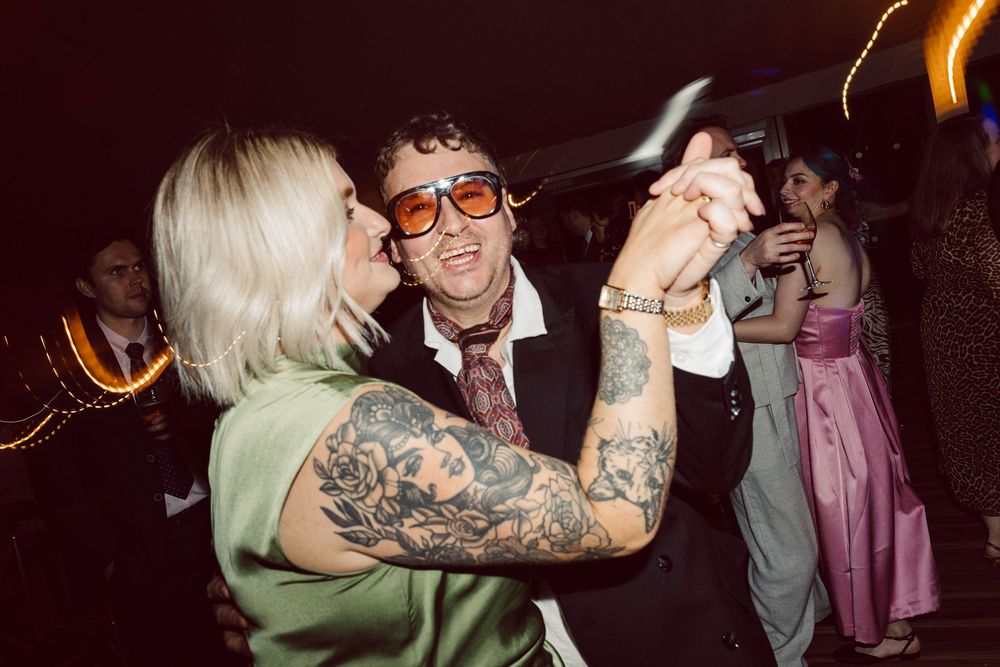 A couple dances together at a party, with one person sporting visible tattoos and the other wearing orange-tinted glasses.