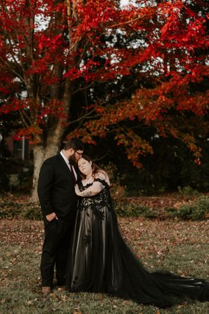 A couple in elegant black attire poses beneath crimson autumn leaves.