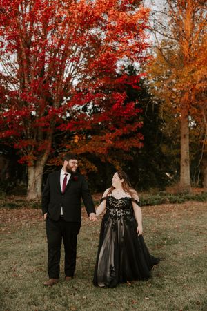 A couple in formal black attire poses together against vibrant autumn foliage with red and orange maple trees.