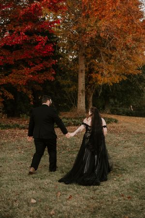 A couple in dark formal attire dances together on a hillside surrounded by vibrant autumn foliage.
