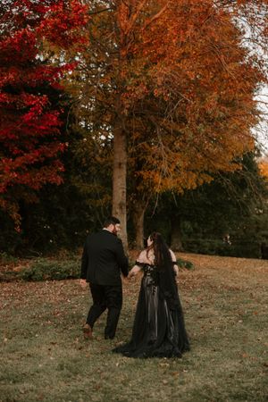 Two people in elegant black clothing share an intimate moment beneath trees with red and orange fall leaves.