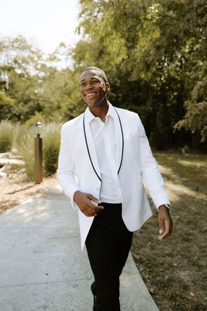 Groom in white jacket and black pants poses confidently in an outdoor setting.