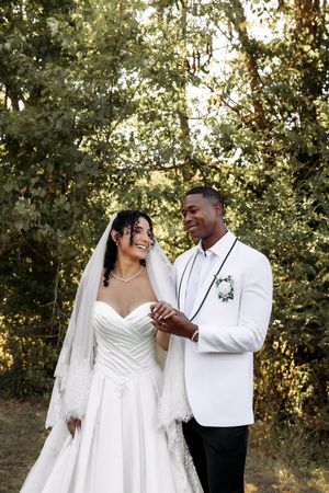A joyful wedding couple sharing a tender moment in their elegant white attire against greenery.