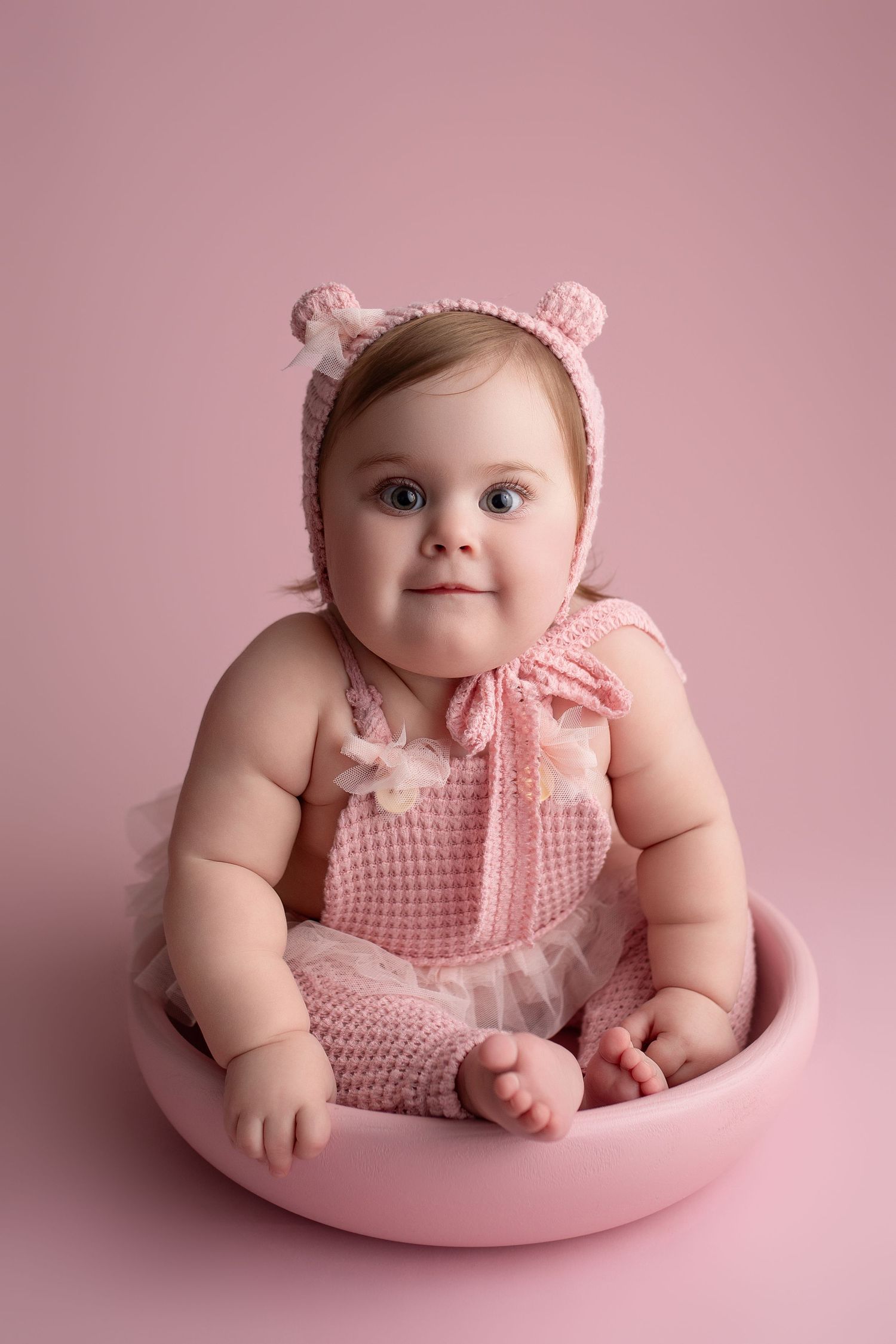 Sweet baby wearing pink knitted outfit with bear ears sits in round pink bowl against pink background.