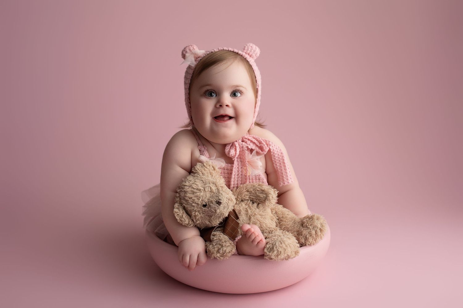 Sweet baby in pink bonnet sits in pink bowl holding teddy bears against rosy background.