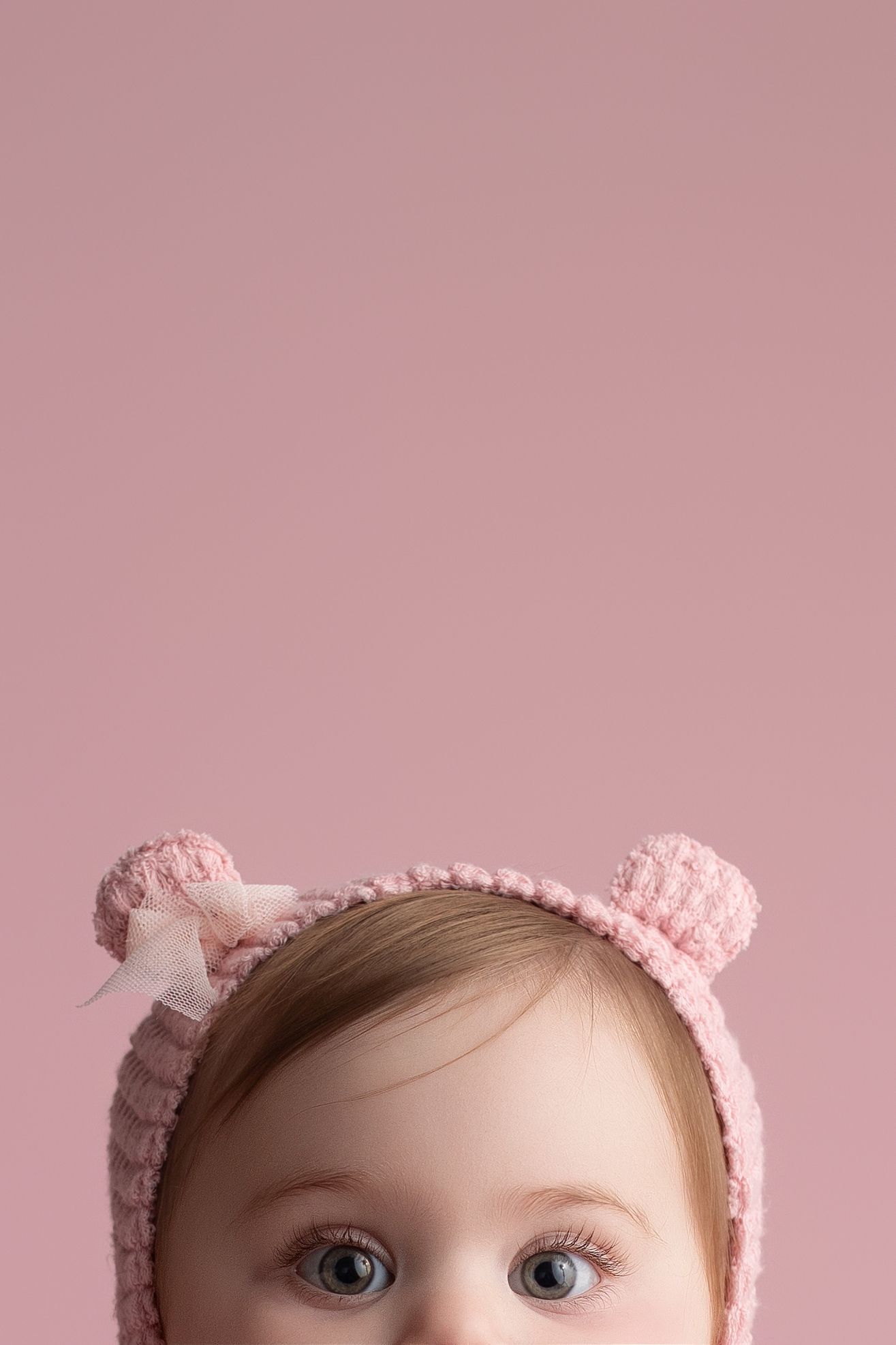 A baby wearing a pink knitted hat with ears against a soft pink background.