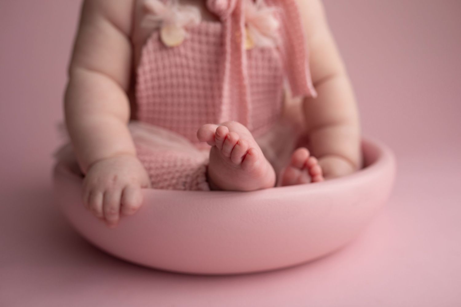 Baby wearing pink gingham romper sits in round pink bowl against pink backdrop during newborn photo session.