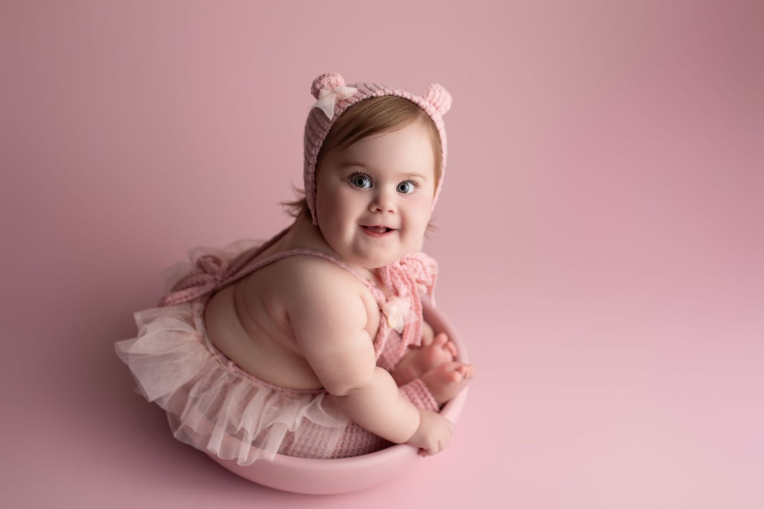A baby wearing a pink tutu and headband lies on a pink backdrop while smiling during a professional photo session.