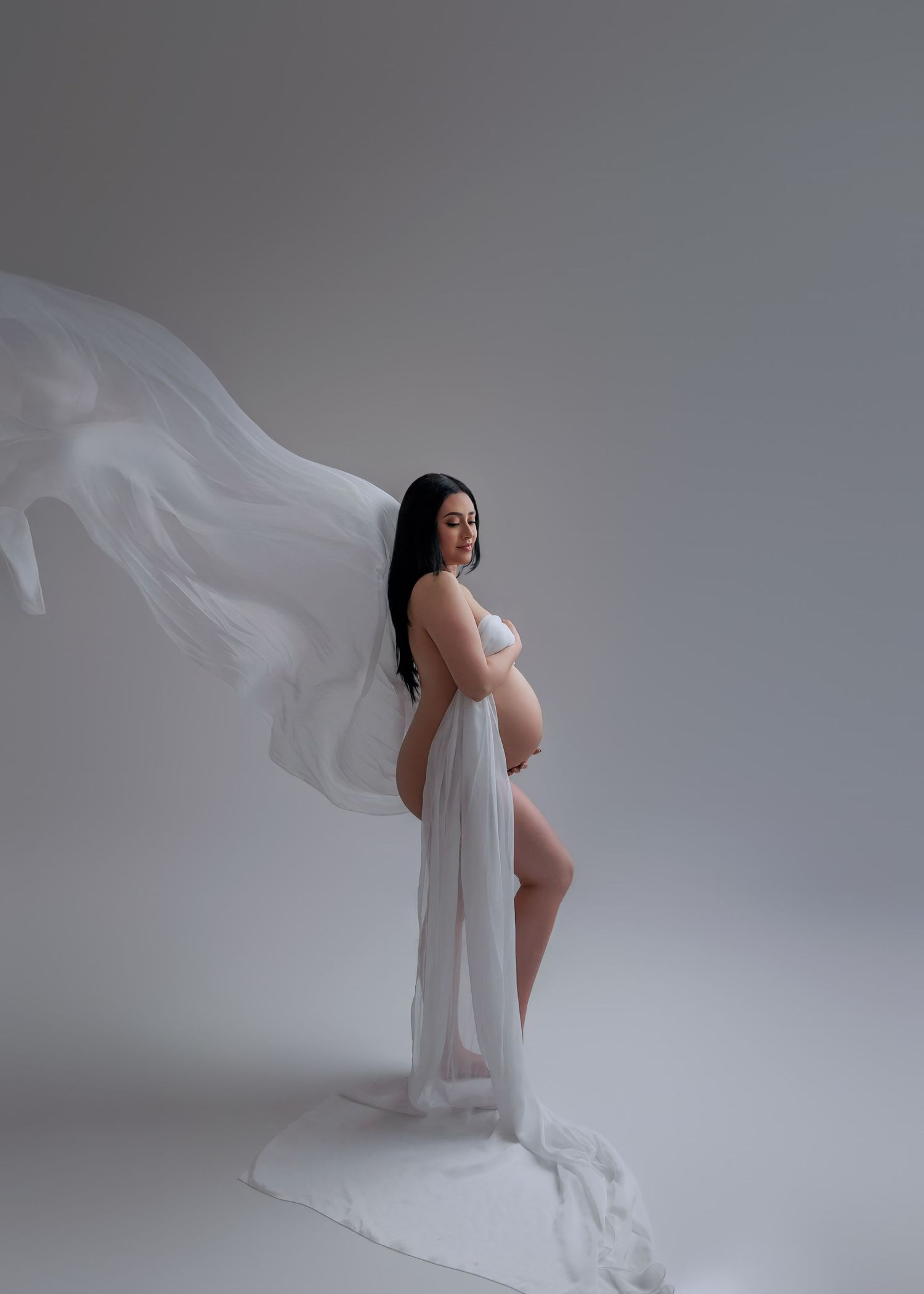 Pregnant woman wrapped in flowing white sheer fabric against a neutral grey background.