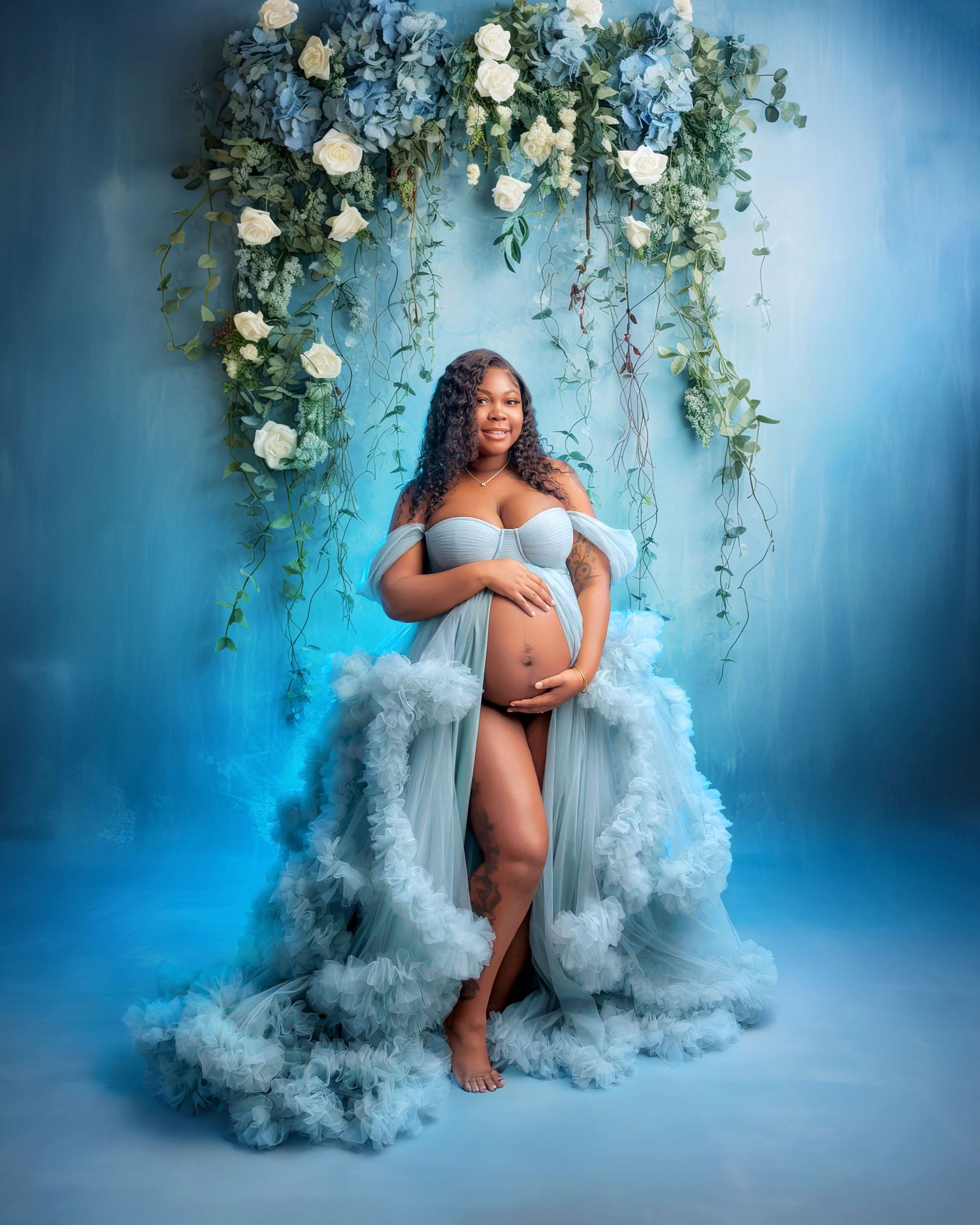 Pregnant woman in a blue ruffled maternity gown poses before a floral arch on a blue studio backdrop.