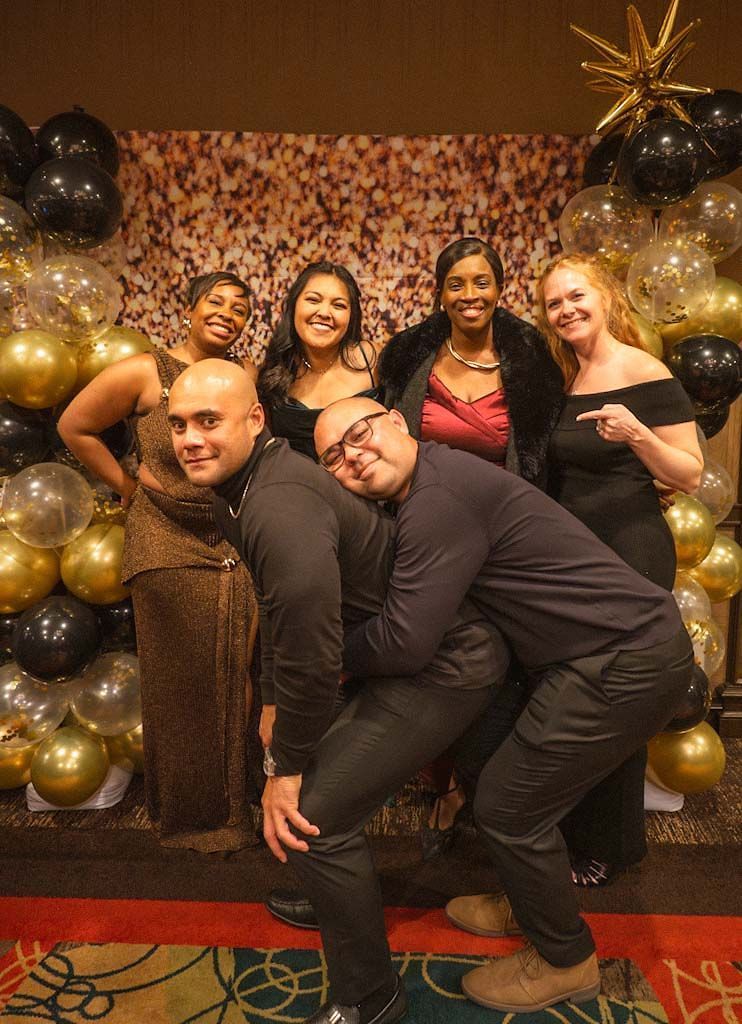 Group of five friends posing playfully in front of gold and black balloon backdrop at festive event.