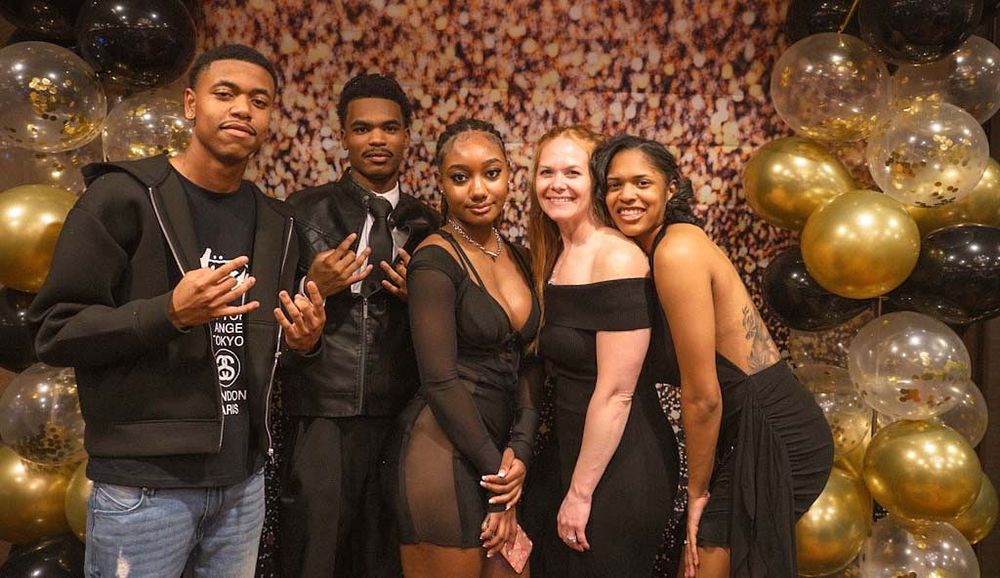 Five young adults smiling together in front of gold and black balloon wall at formal celebration.