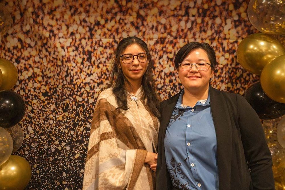 Two women smiling together in front of gold confetti backdrop with black and gold balloons at event.