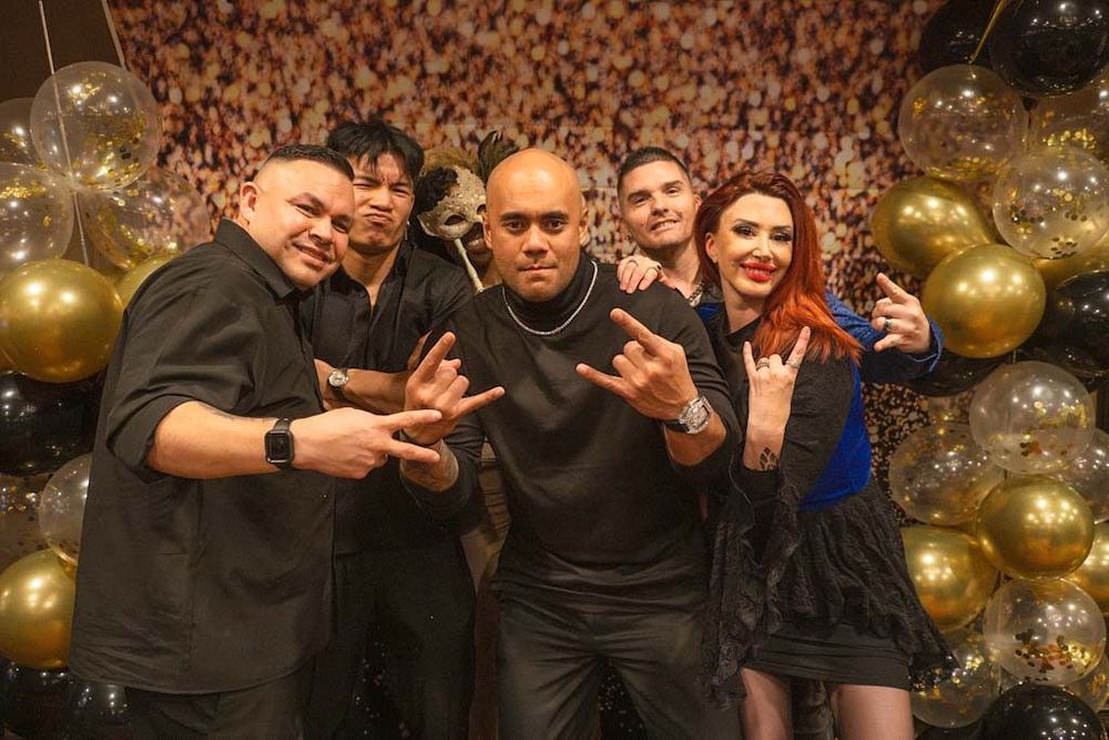 Five people posing with hand gestures in front of gold and black balloon backdrop at party event.