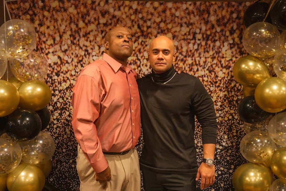 Two men pose together in front of a gold and black balloon arch at a formal celebration event.