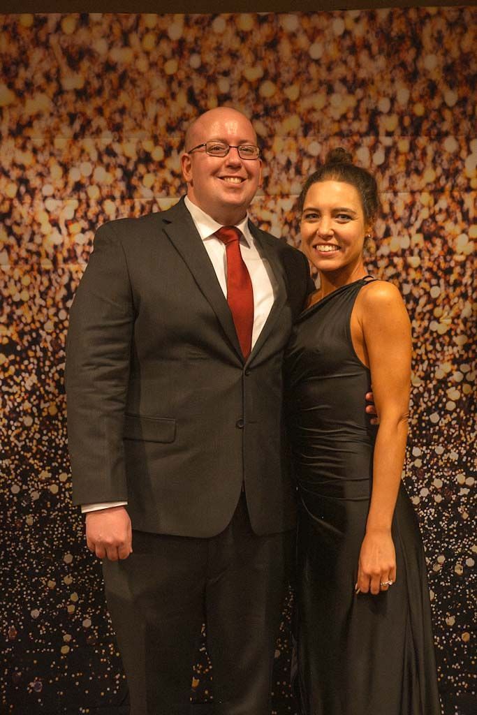 A man in a dark suit with red tie and woman in black dress pose at a formal gala event.