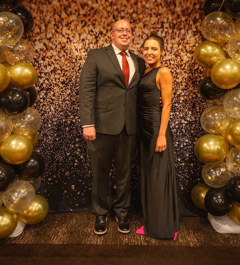 A couple in formal attire poses in front of a gold glitter backdrop with black and gold balloons.