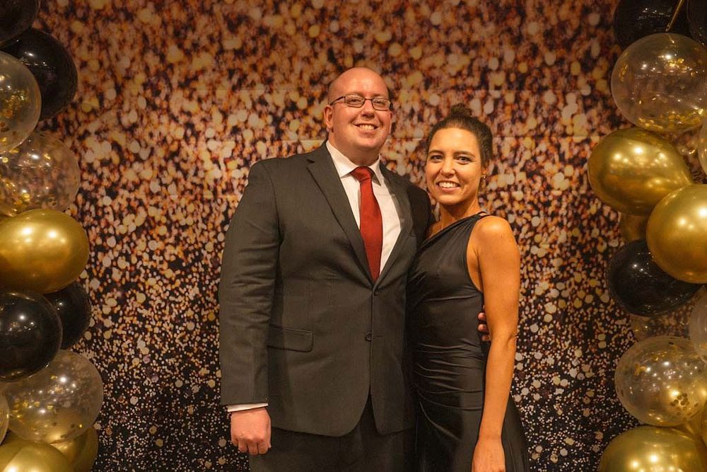 A smiling couple dressed in formal evening wear poses before a sparkling gold balloon backdrop.