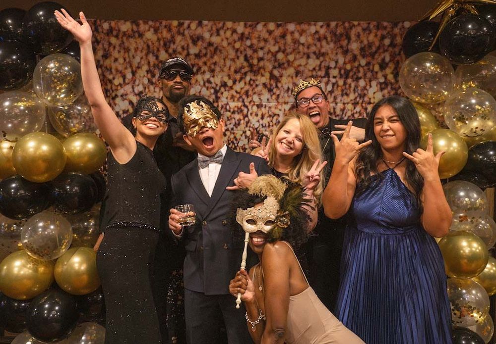 A lively group of five people in elegant attire pose with masquerade masks at a gala.