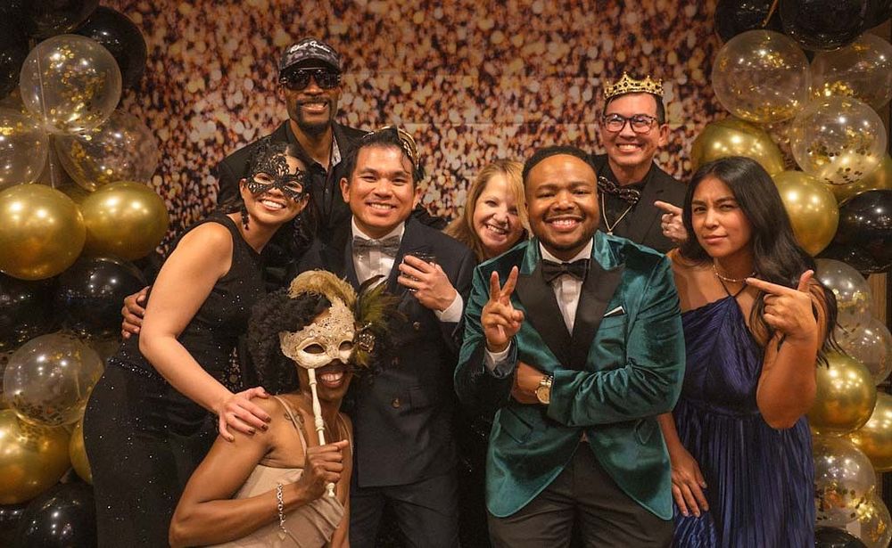 A group of seven people in formal and masquerade attire pose with gold and black balloons.