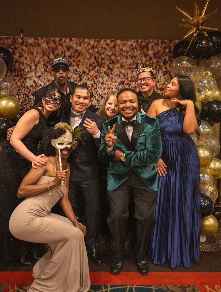 A group of eight guests in formal masquerade attire pose joyfully at a glamorous gala event.