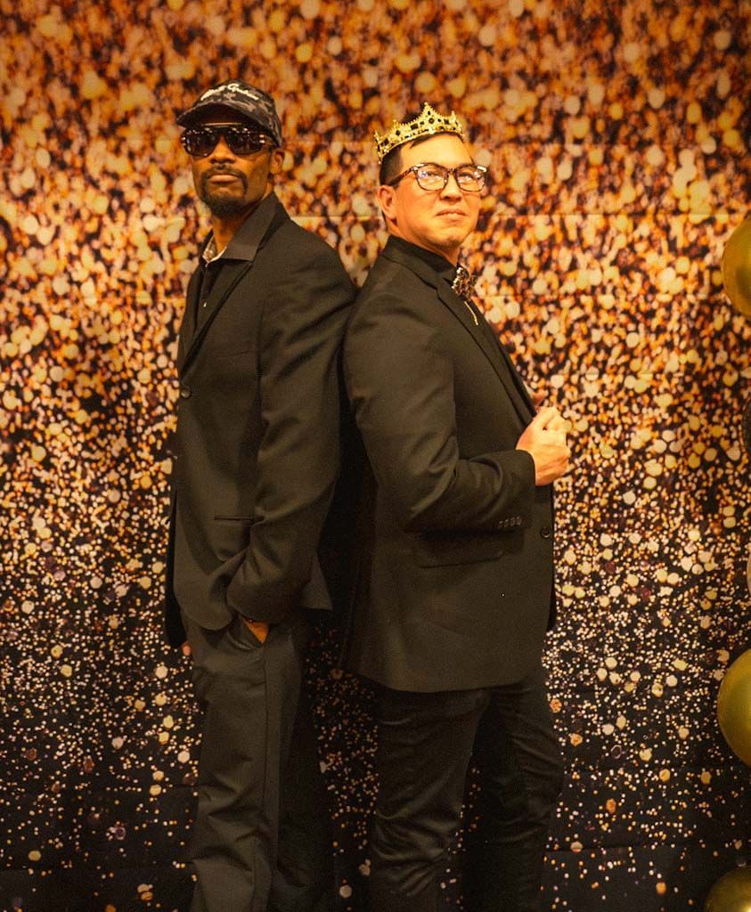 Two men dressed in all-black formal suits pose back-to-back before a gold glitter backdrop.