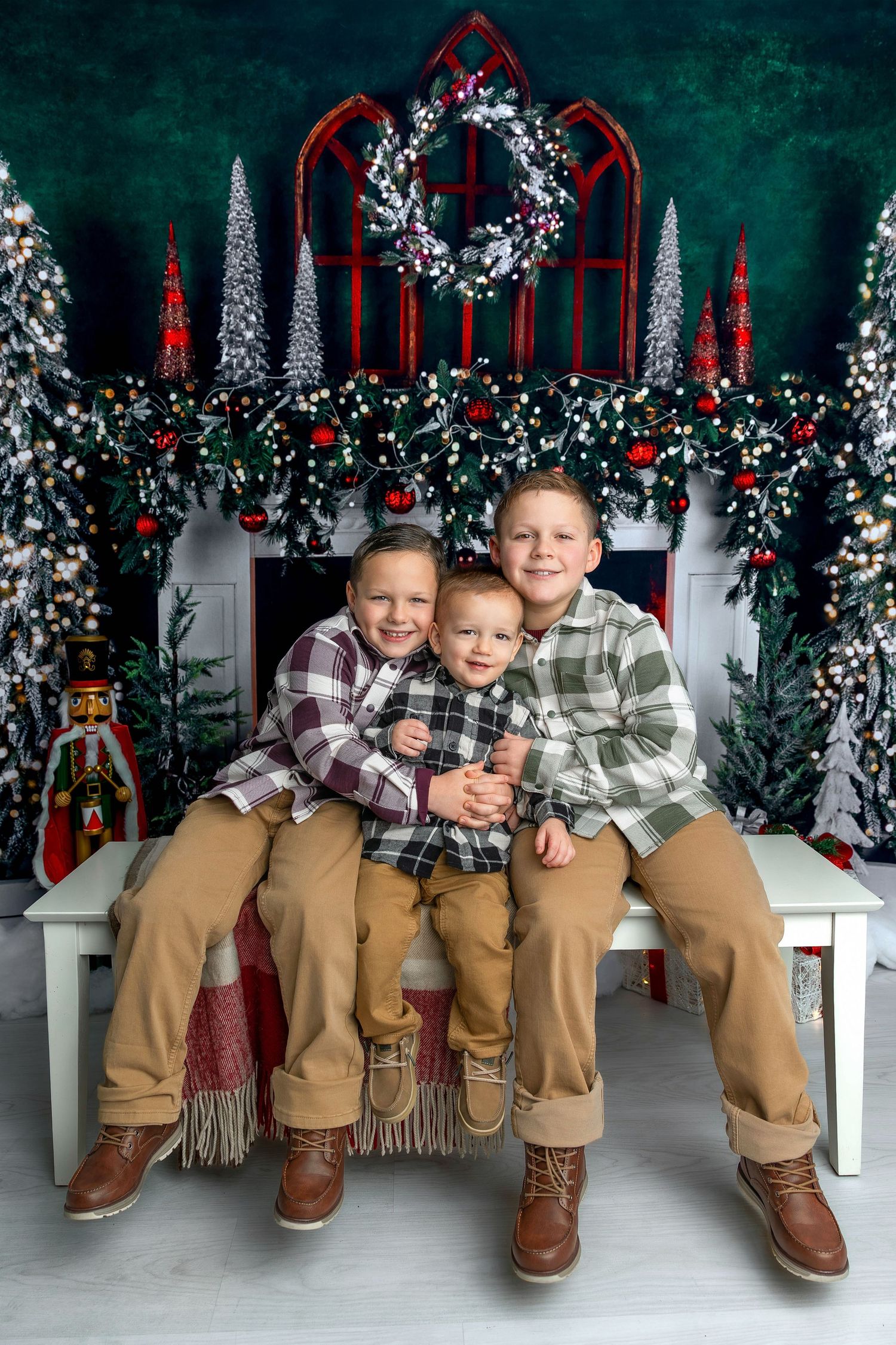 Four children in matching plaid shirts and khaki pants sit on a bench against a festive Christmas backdrop with wreaths.