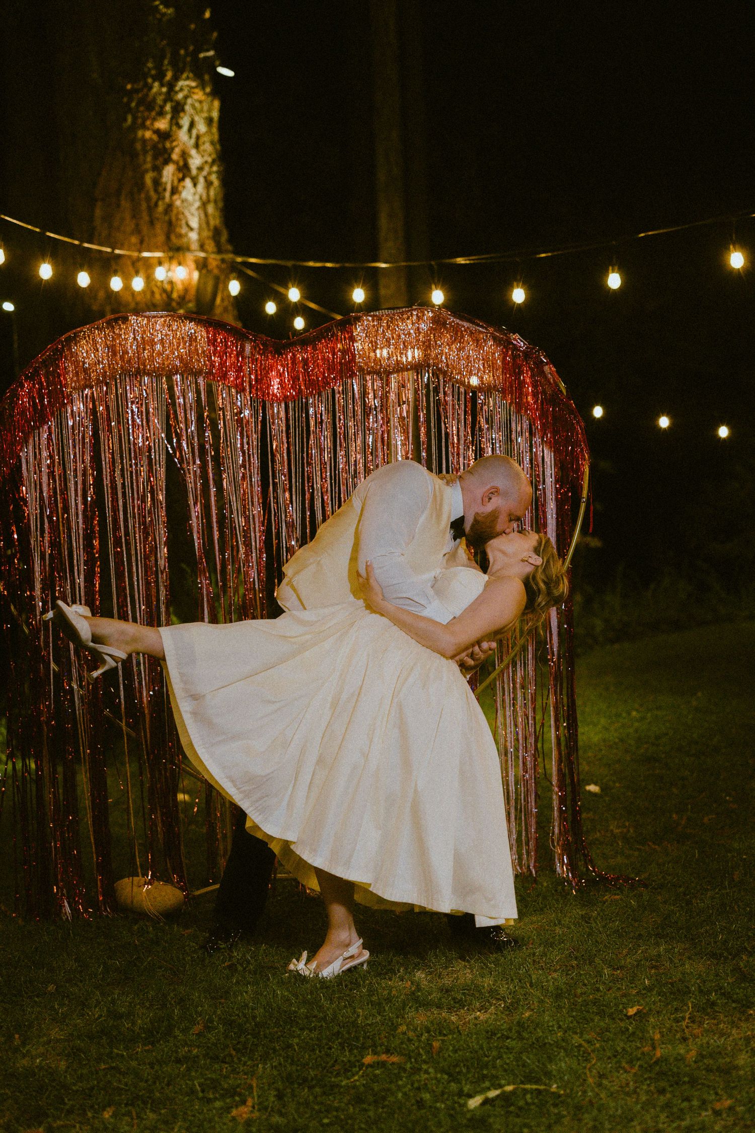 A person in a white dress being dramatically dipped and kissed by their newly wed spouse against a heart-shaped red tinsel backdrop.