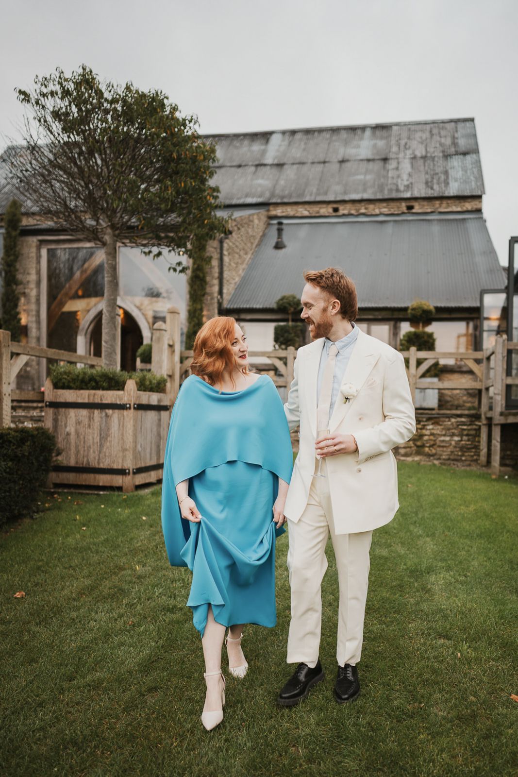 A couple in light colored outfits embrace while walking on grass with rustic farm buildings in the background.