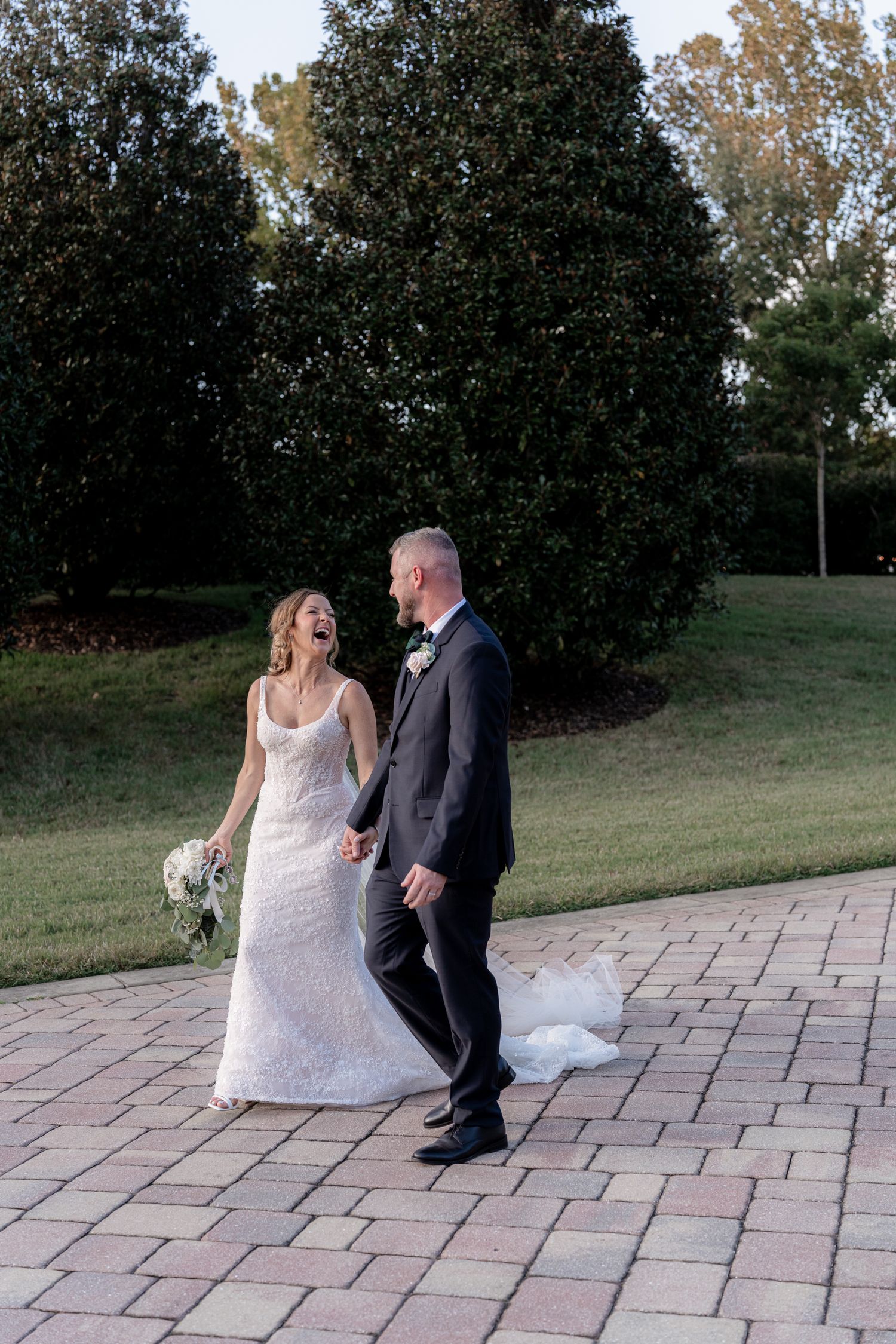 A couple in wedding attire embraces in a garden setting with yellow flowering bushes and olive trees in soft sunlight.