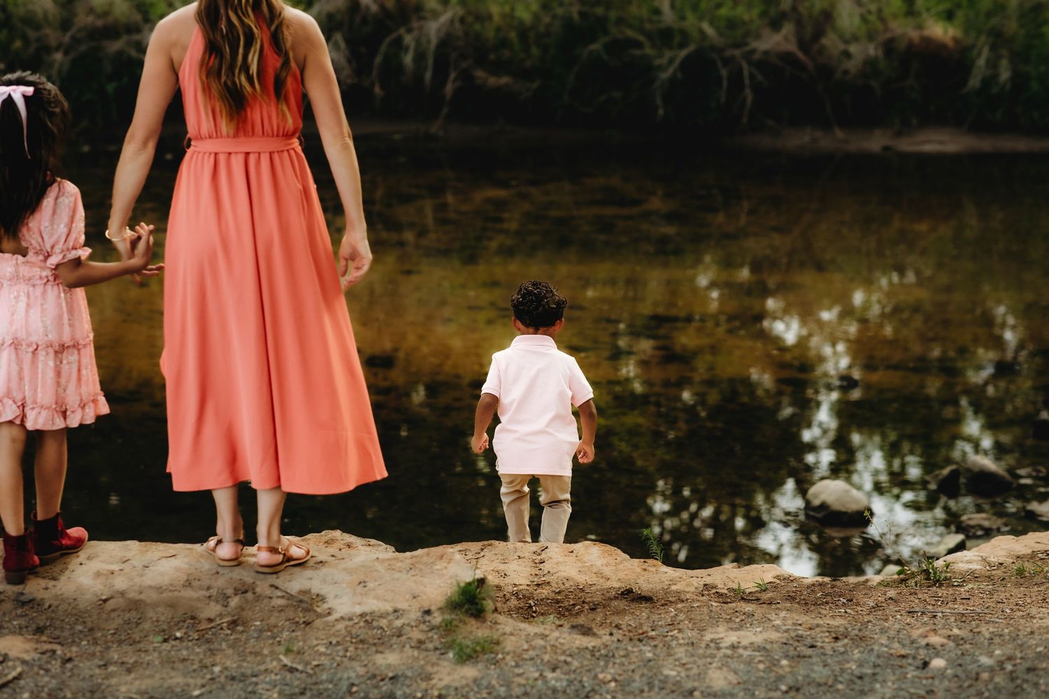 A group in coral pink dresses and formal attire stands near a reflective pond in an outdoor setting.