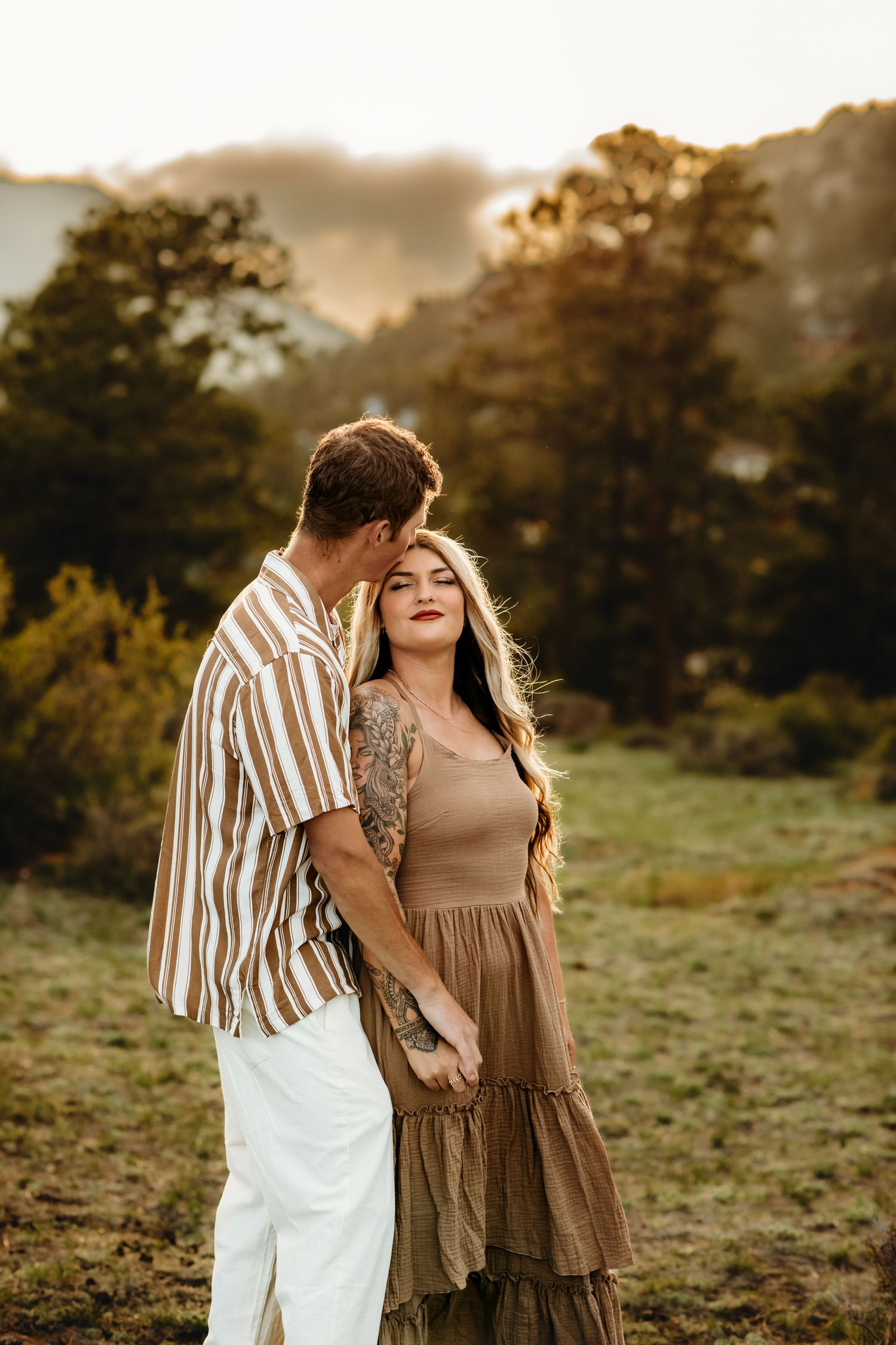 A couple embraces in a sunlit meadow at golden hour wearing brown and striped outfits.