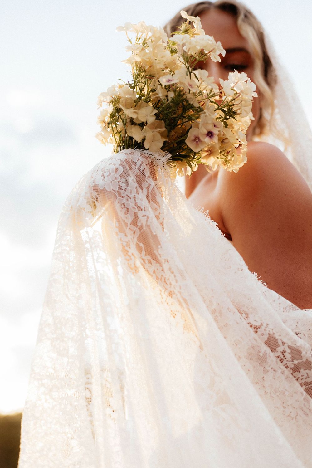 Soft focus detail of white lace wedding dress and cream-colored floral bouquet in natural light.