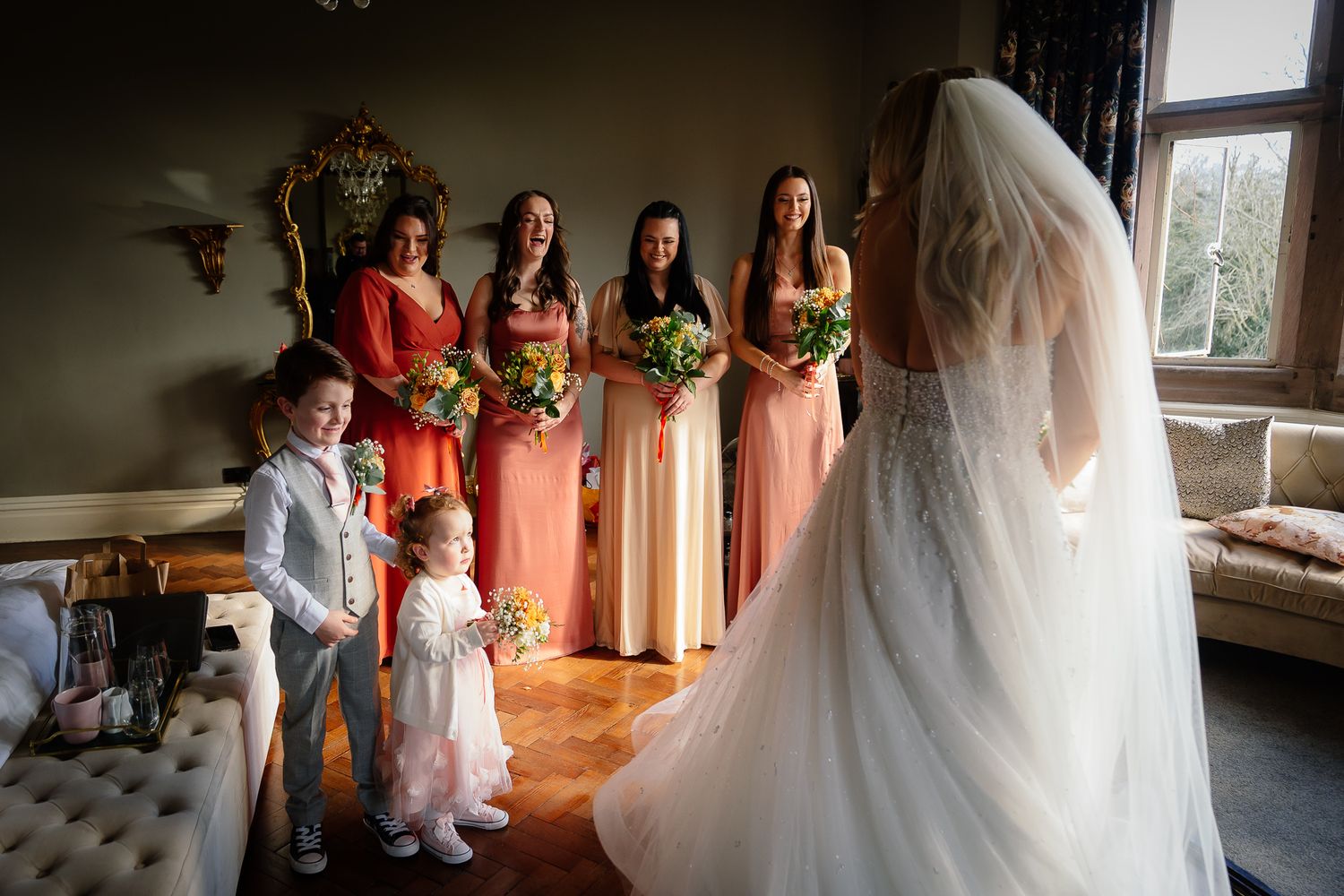 Bridesmaids in coral dresses gather by a window during wedding preparations.