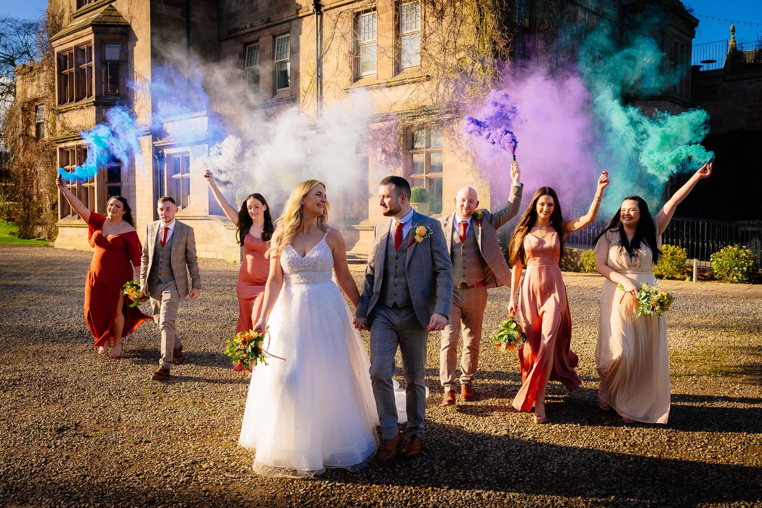 Wedding party poses with colorful smoke bombs outside historic building.