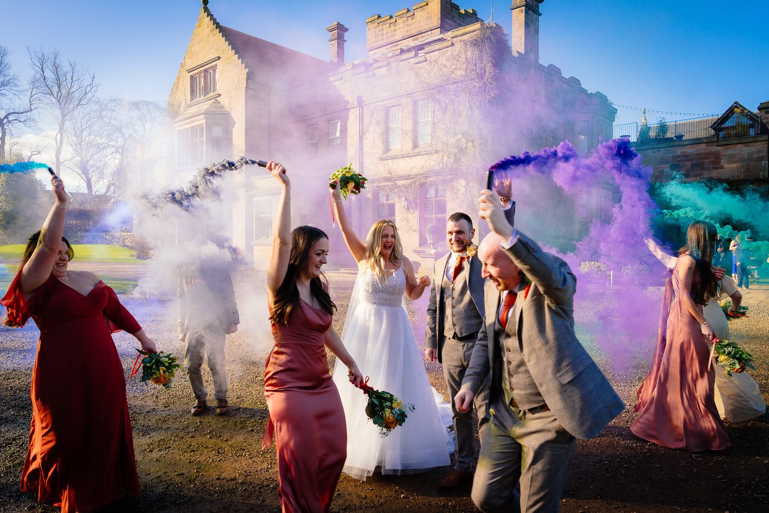 Wedding group creates dramatic scene with purple smoke effects against blue sky.