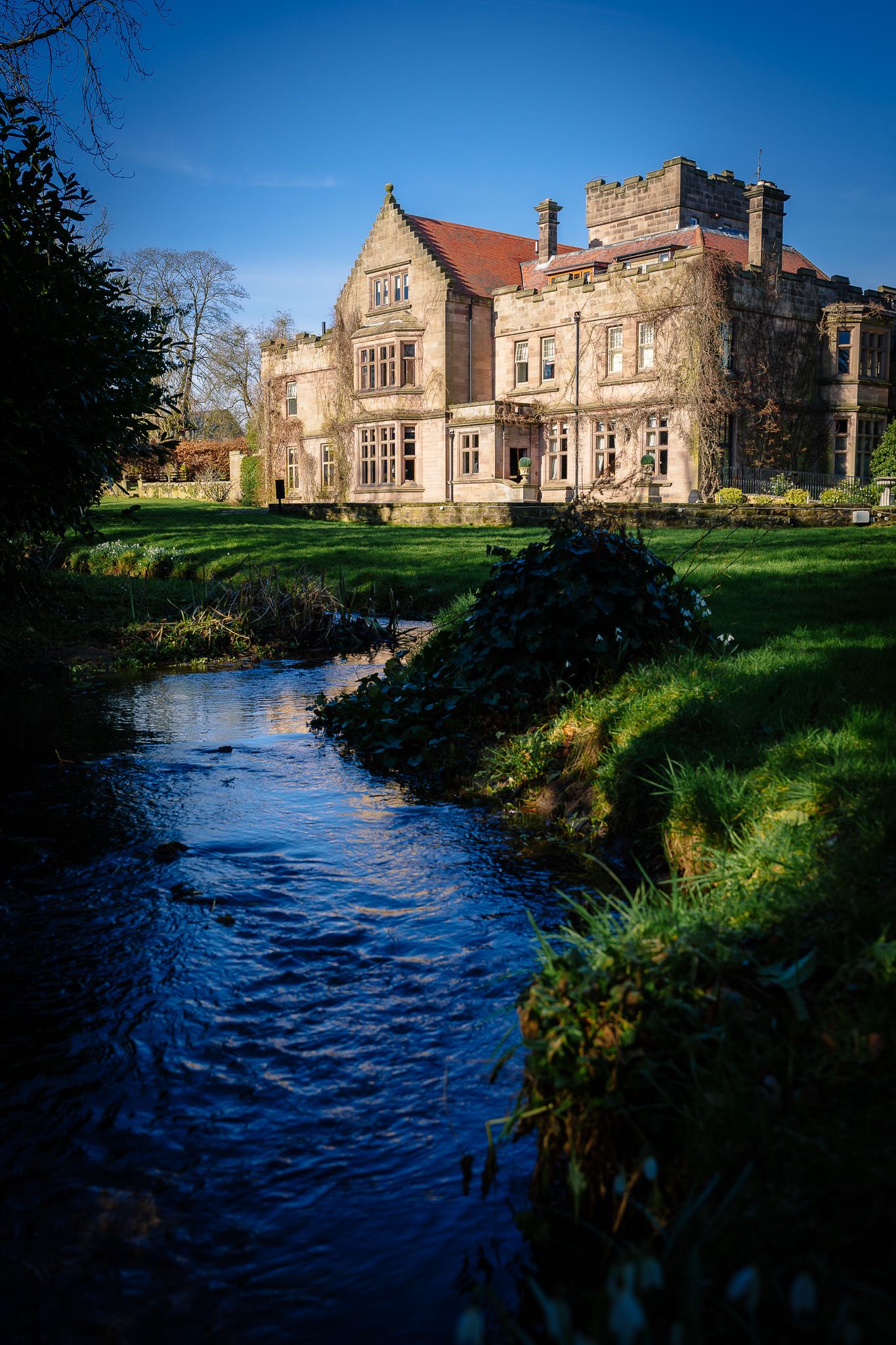 Historic stone manor house beside a flowing stream under bright blue sky.
