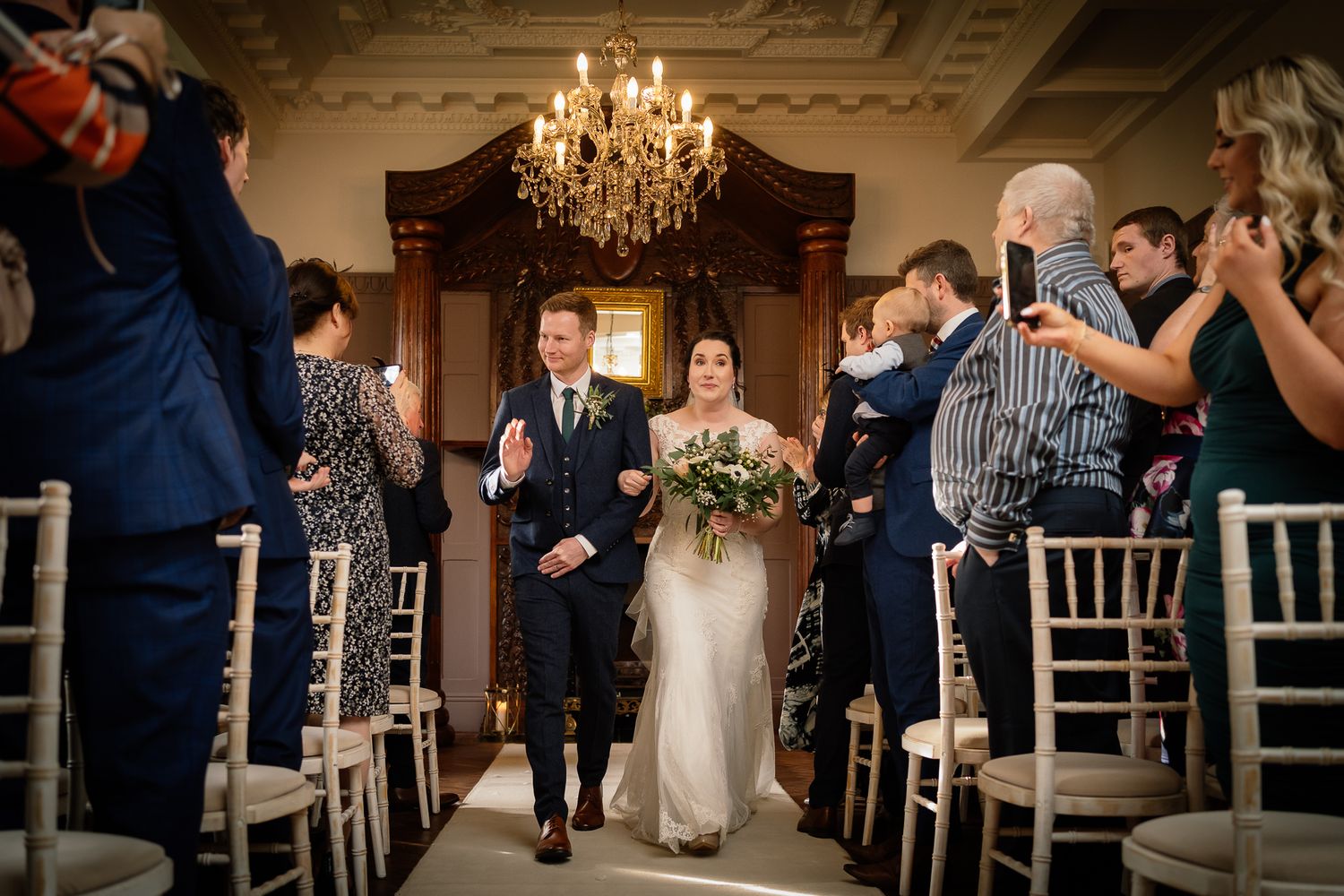 Wedding reception venue with elegant chandelier and guests seated at decorated tables. Ellingham Hall