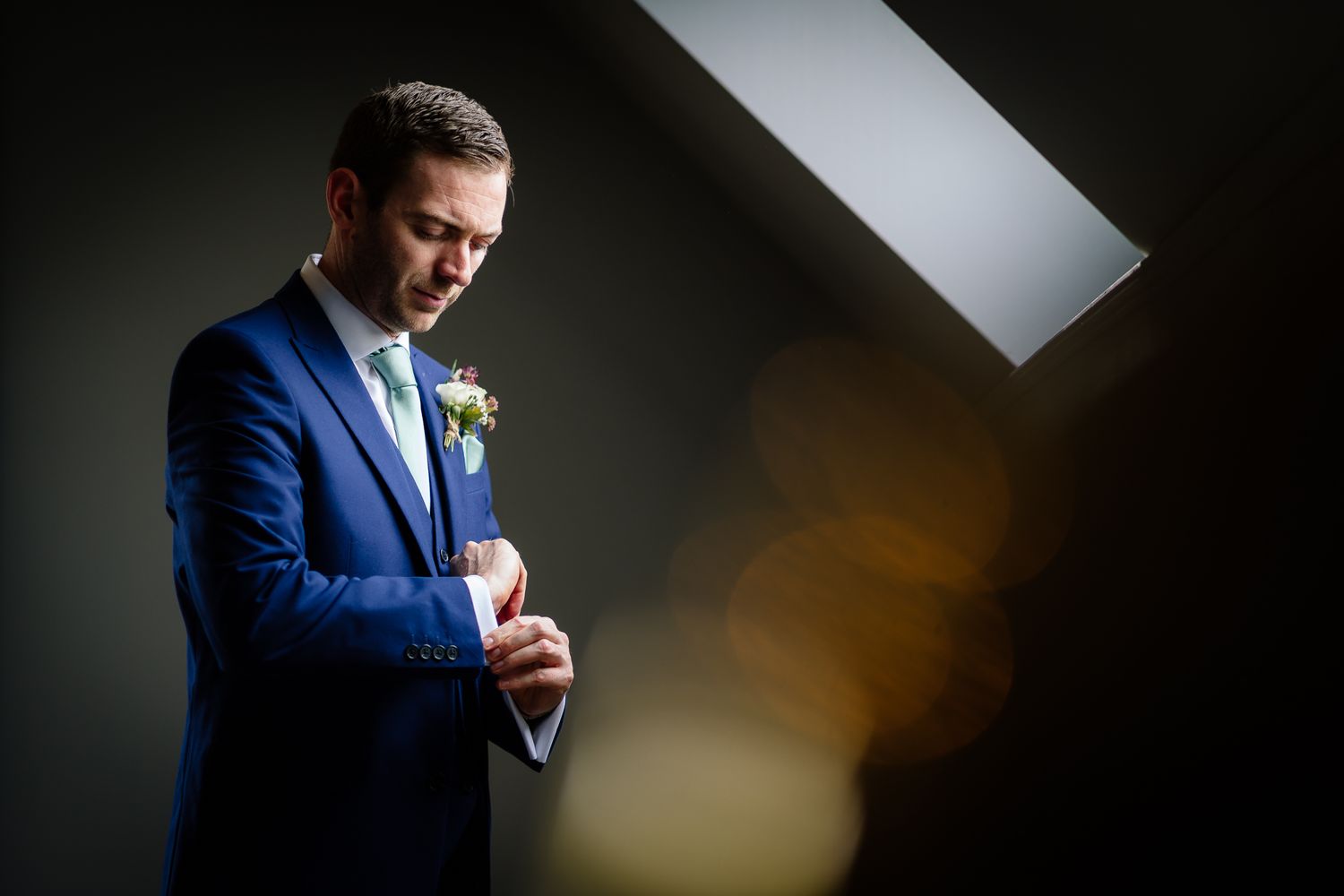 Dramatic lighting illuminates a groom adjusting his tie in a navy suit.