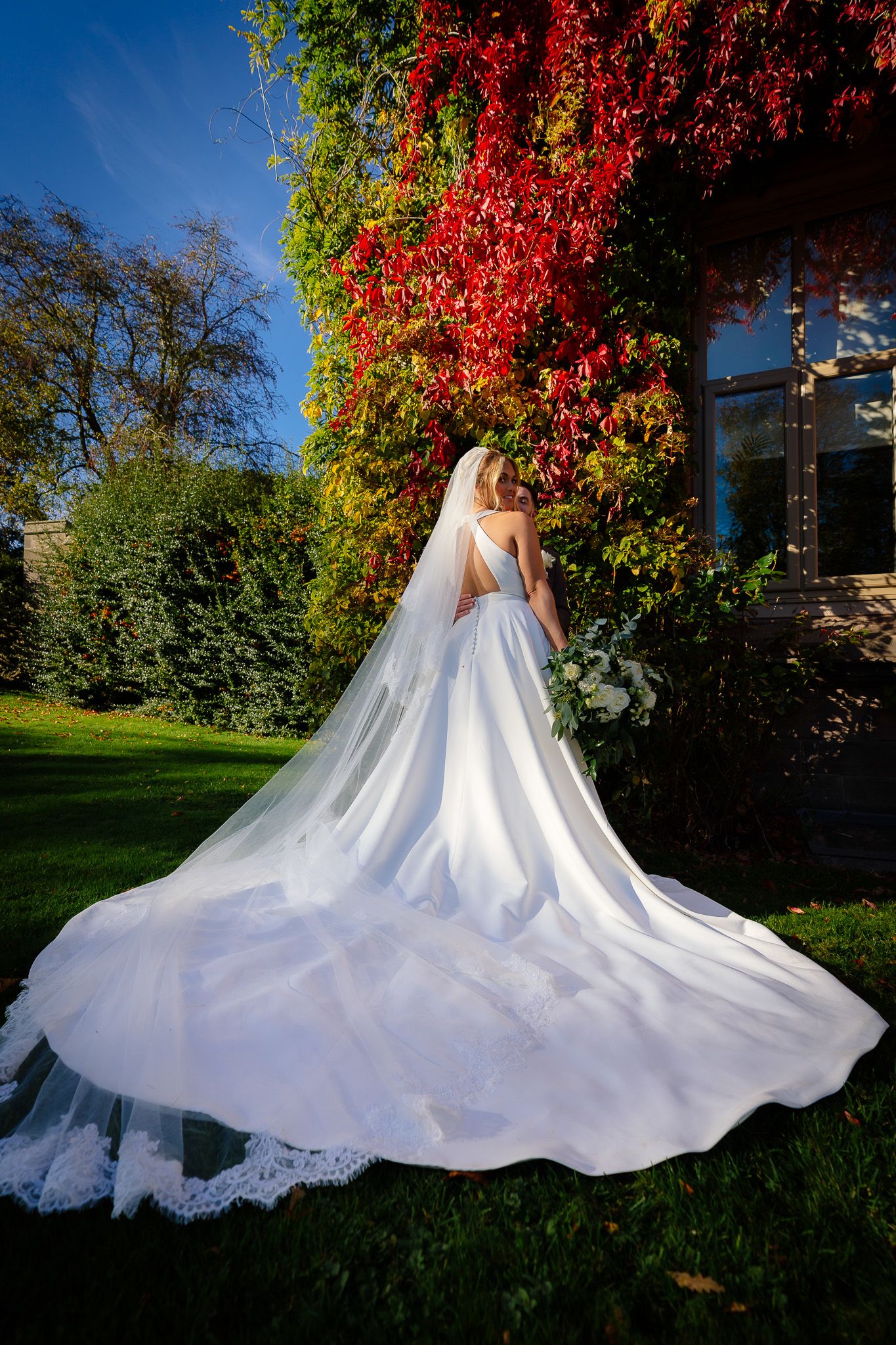 A flowing white wedding dress with cathedral train spreads across green grass beside a wall covered in red autumn ivy.