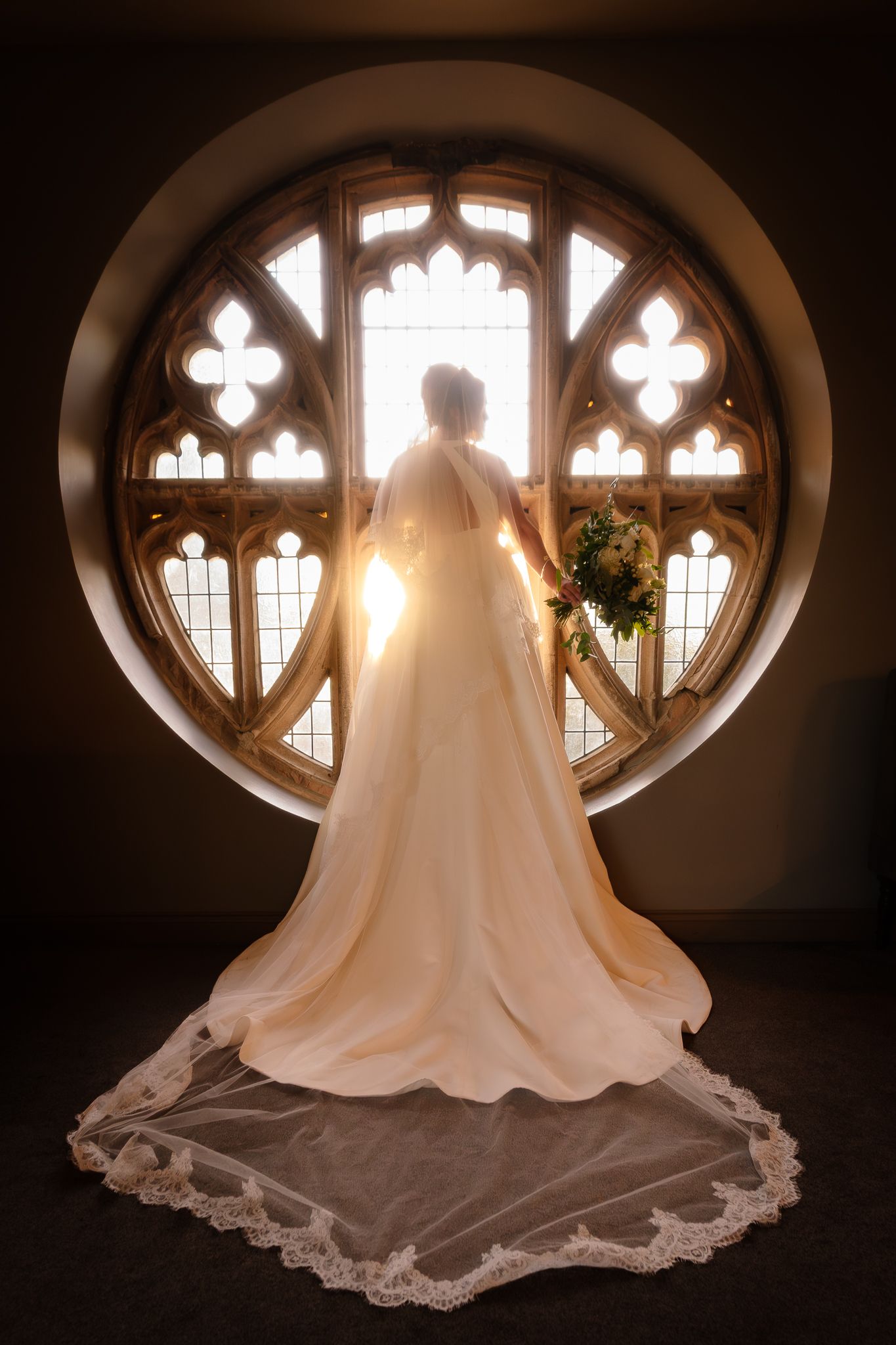 A wedding dress silhouette stands before an ornate gothic window with dramatic backlighting.