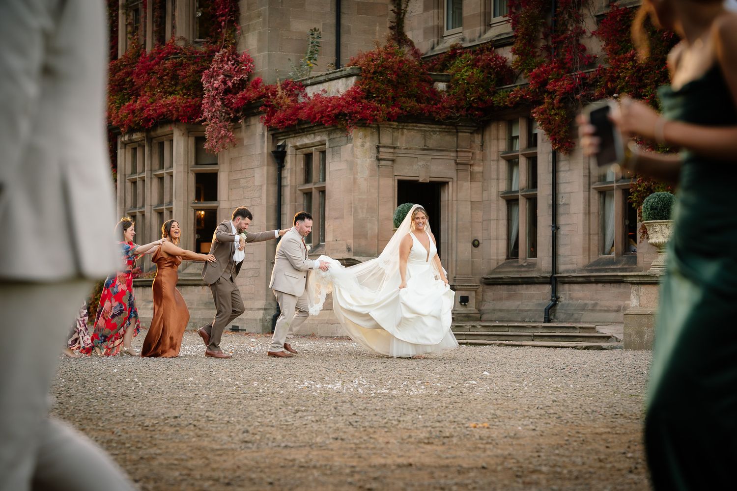 Wedding party runs playfully across a courtyard with red ivy-covered buildings in the background.