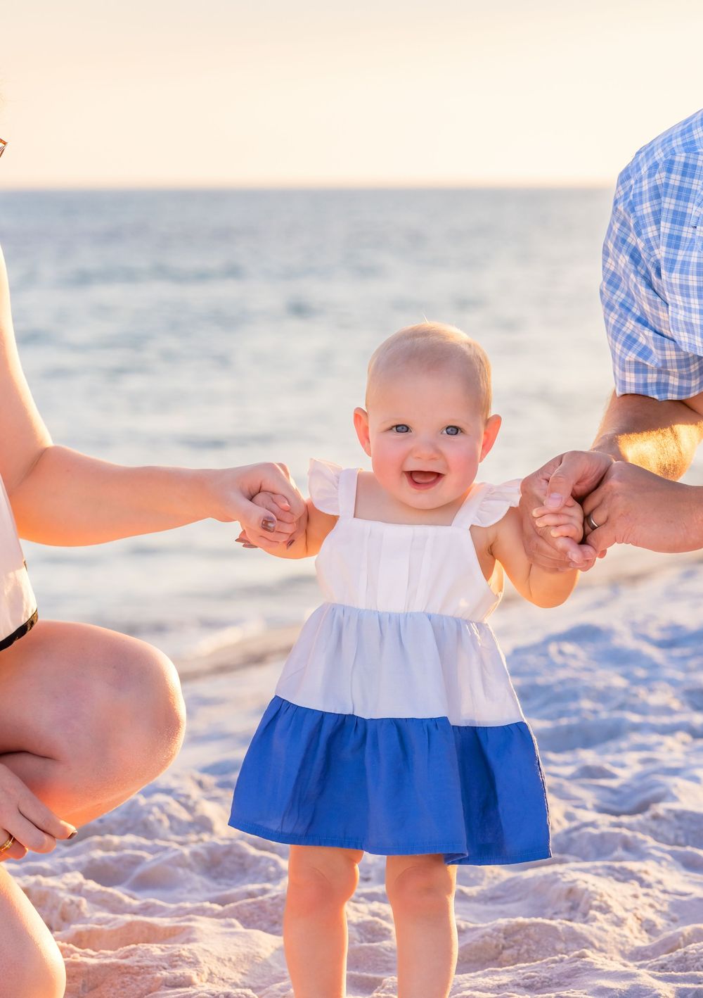 A baby in a white and blue dress smiles while holding hands on a sunny beach at sunset.