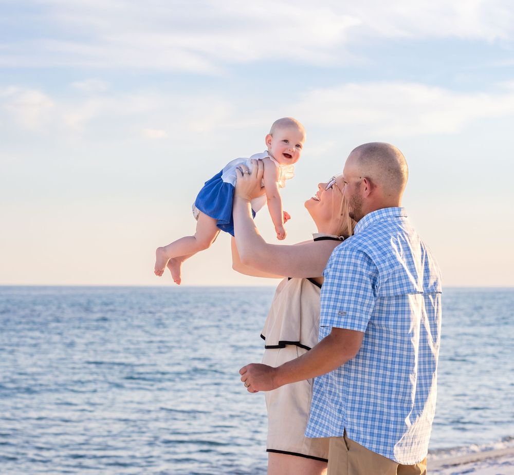 A joyful family moment by the ocean at sunset with a baby being lifted into the air against a blue sky backdrop.