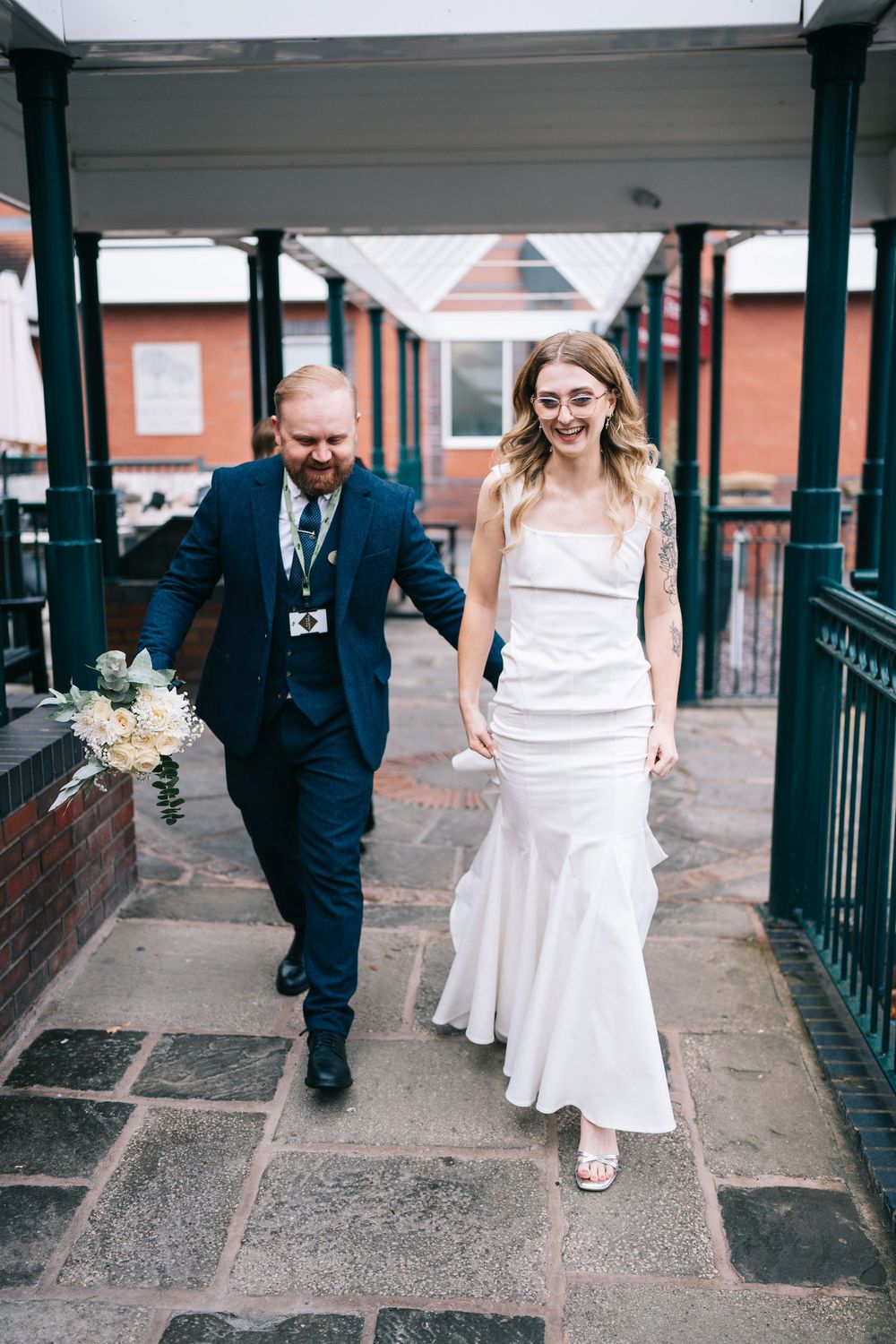 A wedding couple walks along a covered brick pathway with green pillars on a cloudy day.