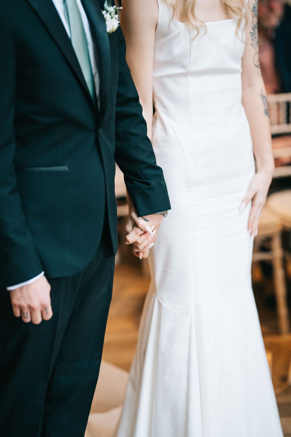 Close-up details of a bride in white gown holding hands with groom in black tuxedo at wedding ceremony.