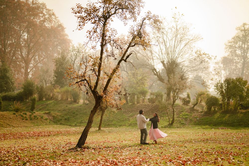 Couple walking hand-in-hand through a misty autumn park scattered with fallen leaves beside a bare tree.