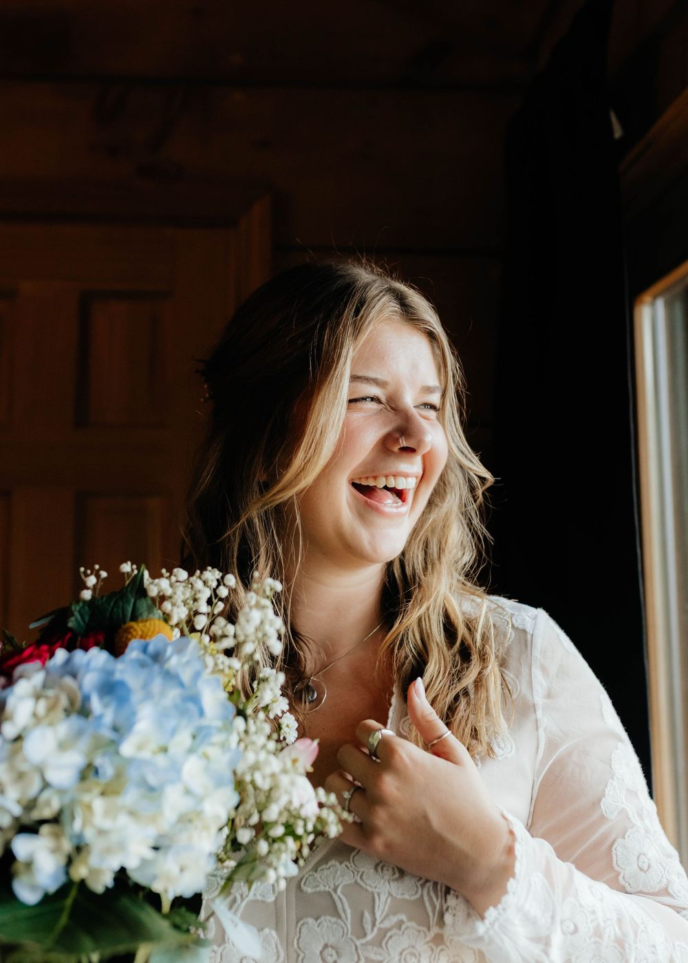 A person in a white lace dress laughs joyfully while holding delicate white baby's breath flowers near a window.