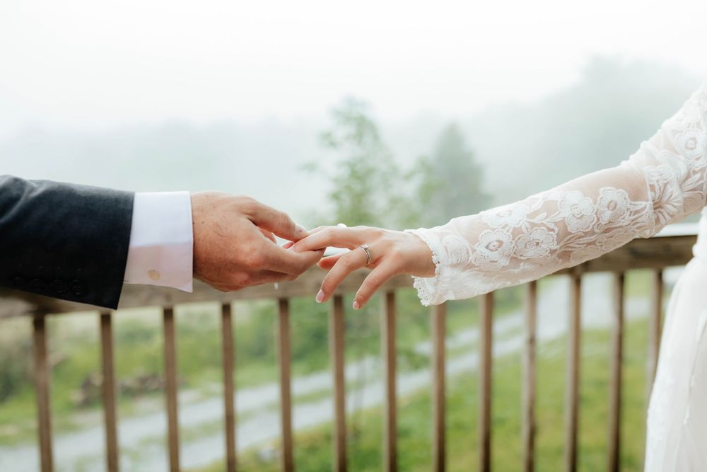 Close-up of hands exchanging wedding rings against a misty mountain backdrop with white lace sleeve detail.