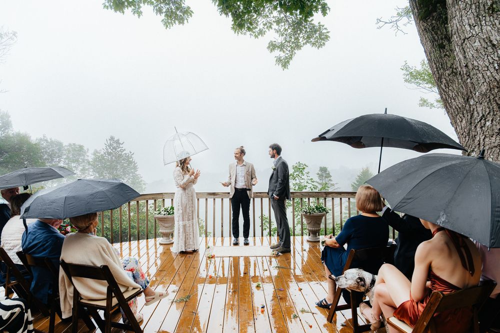 Wedding guests gather under umbrellas on a rainy day at an outdoor venue with white railings.
